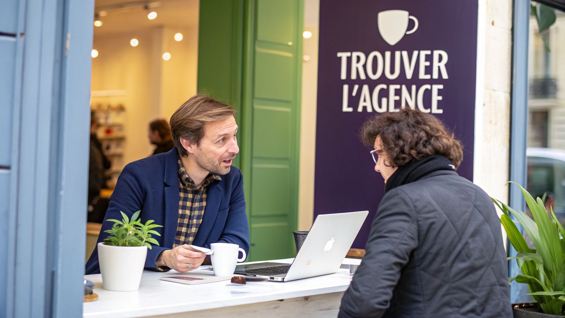Deux hommes discutent sérieusement devant un ordinateur portable, avec café et plante verte. L'un explique, l'autre écoute.