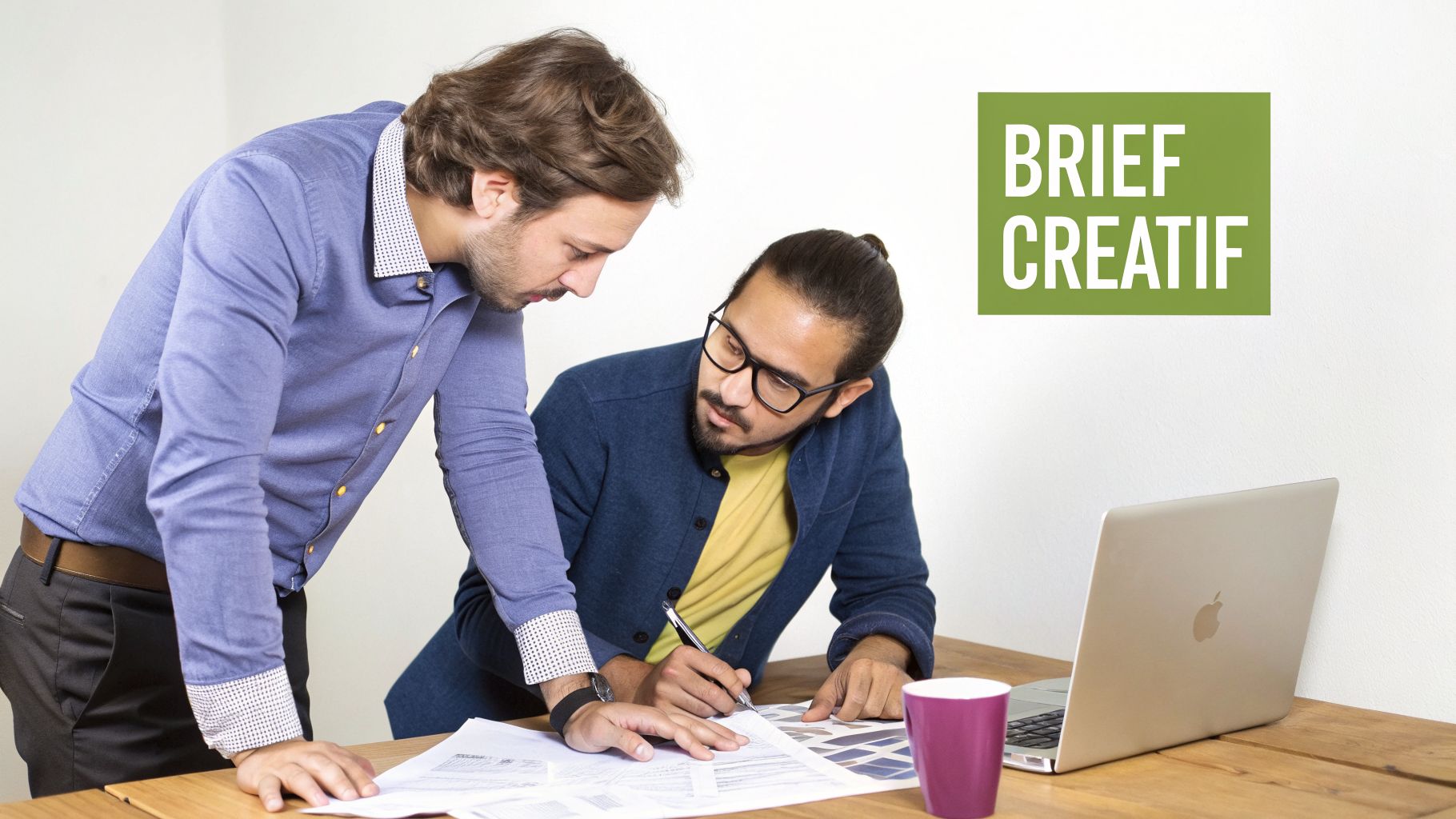 Deux hommes discutent d'un brief créatif, examinant des documents et des échantillons de couleurs sur un bureau.