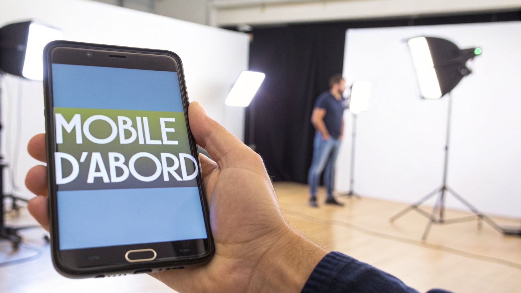 Main tenant un smartphone affichant 'MOBILE D'ABORD' dans un studio photo avec un homme flou et des lumières.